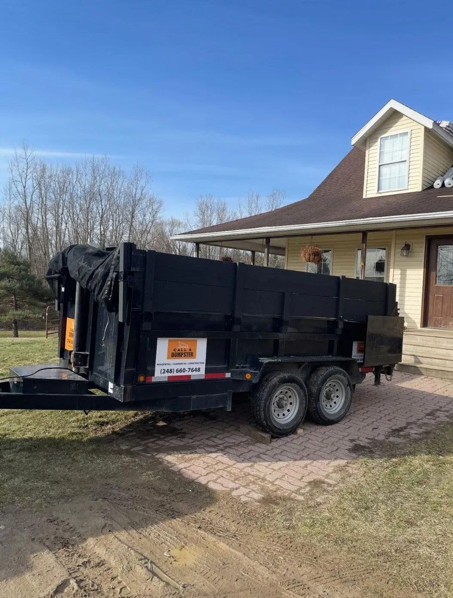 Black dumpster trailer parked in front of a light yellow house with a blue sky.