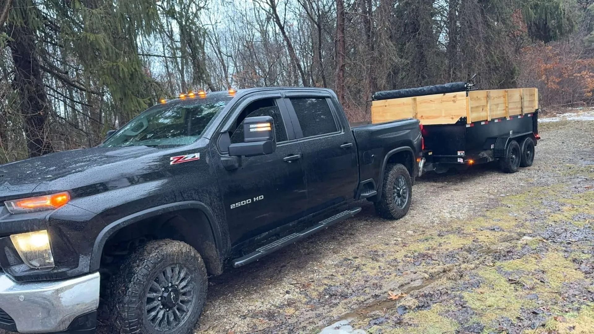 Black pickup truck towing a trailer on a muddy, snowy road in a wooded area.