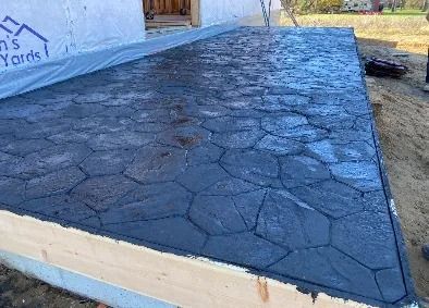 Freshly poured, dark gray concrete patio with textured, stone-like pattern; wood border.