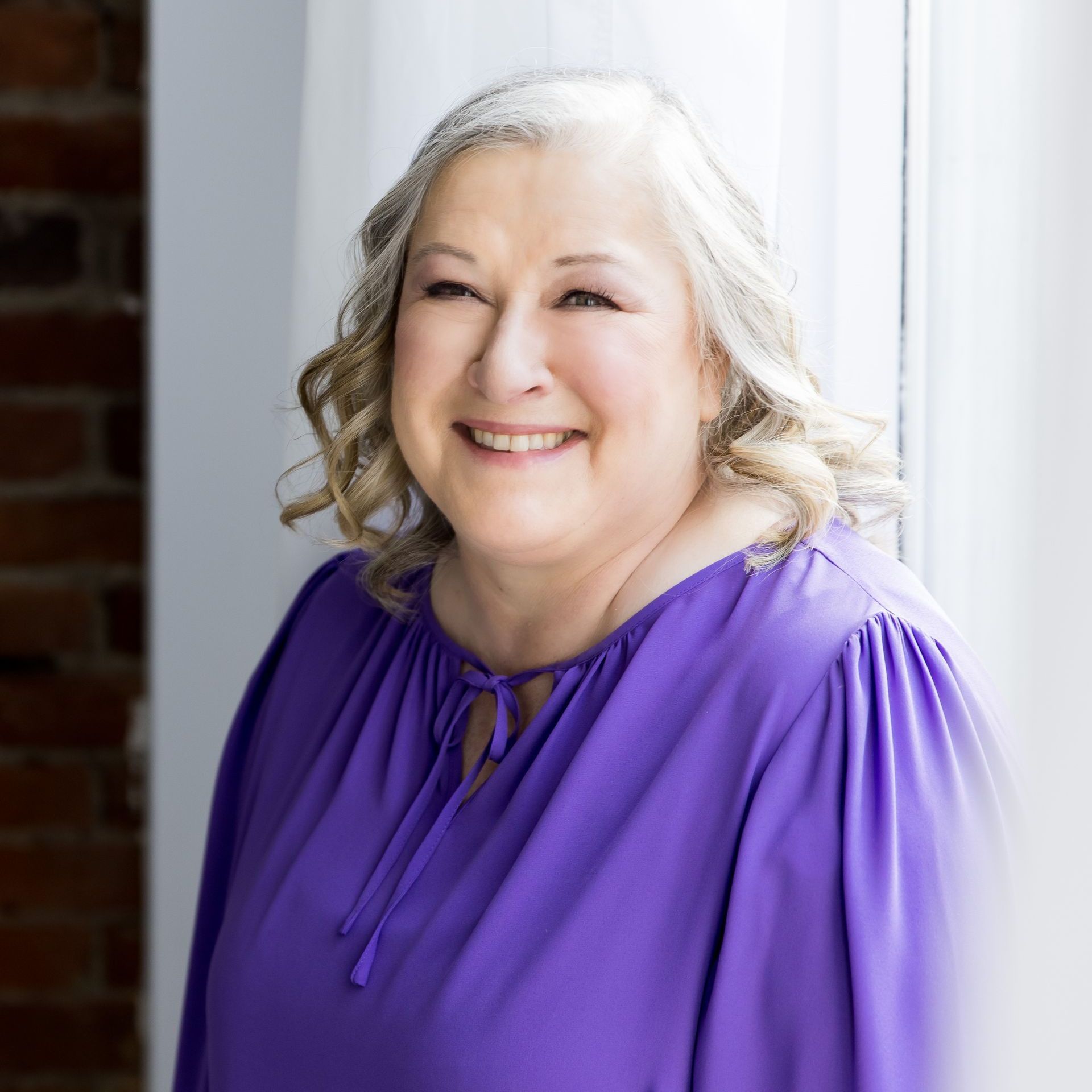 A woman in a purple shirt is smiling and standing next to a window.