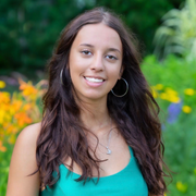 Smiling woman in a green top standing outdoors in a garden with colorful flowers.