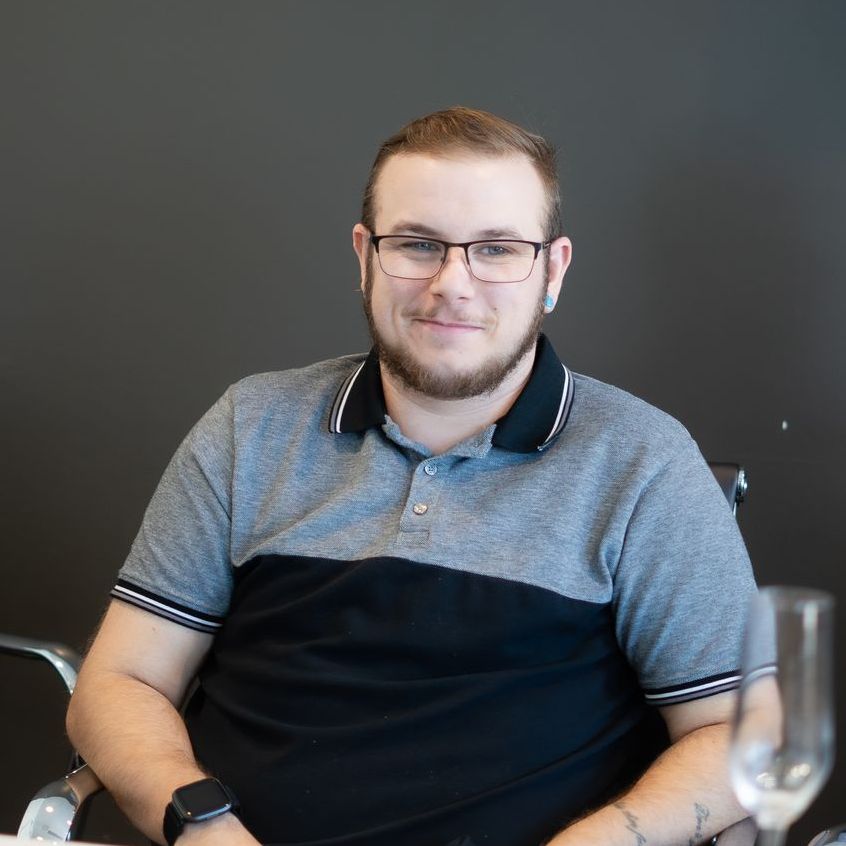 A man with glasses and a beard is sitting at a table with a glass of wine.