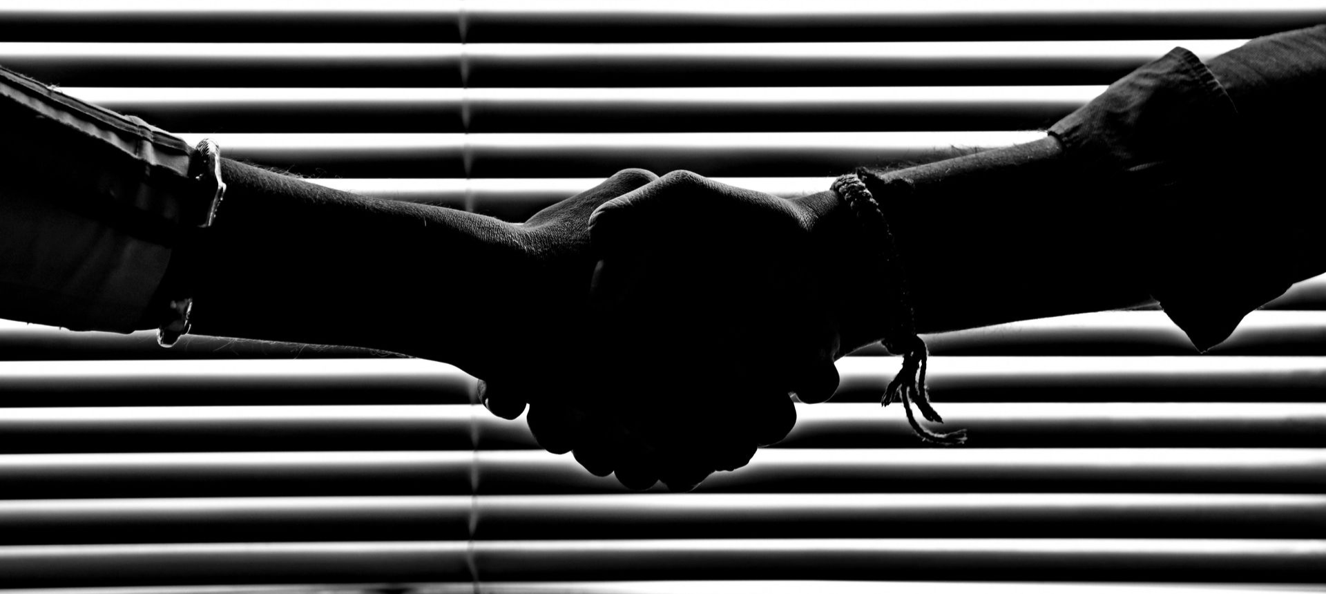 Silhouette of two hands shaking in front of horizontal blinds, suggesting agreement or a business deal.
