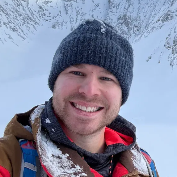 Smiling person in a dark knit hat and winter jacket, standing in a snowy mountain landscape