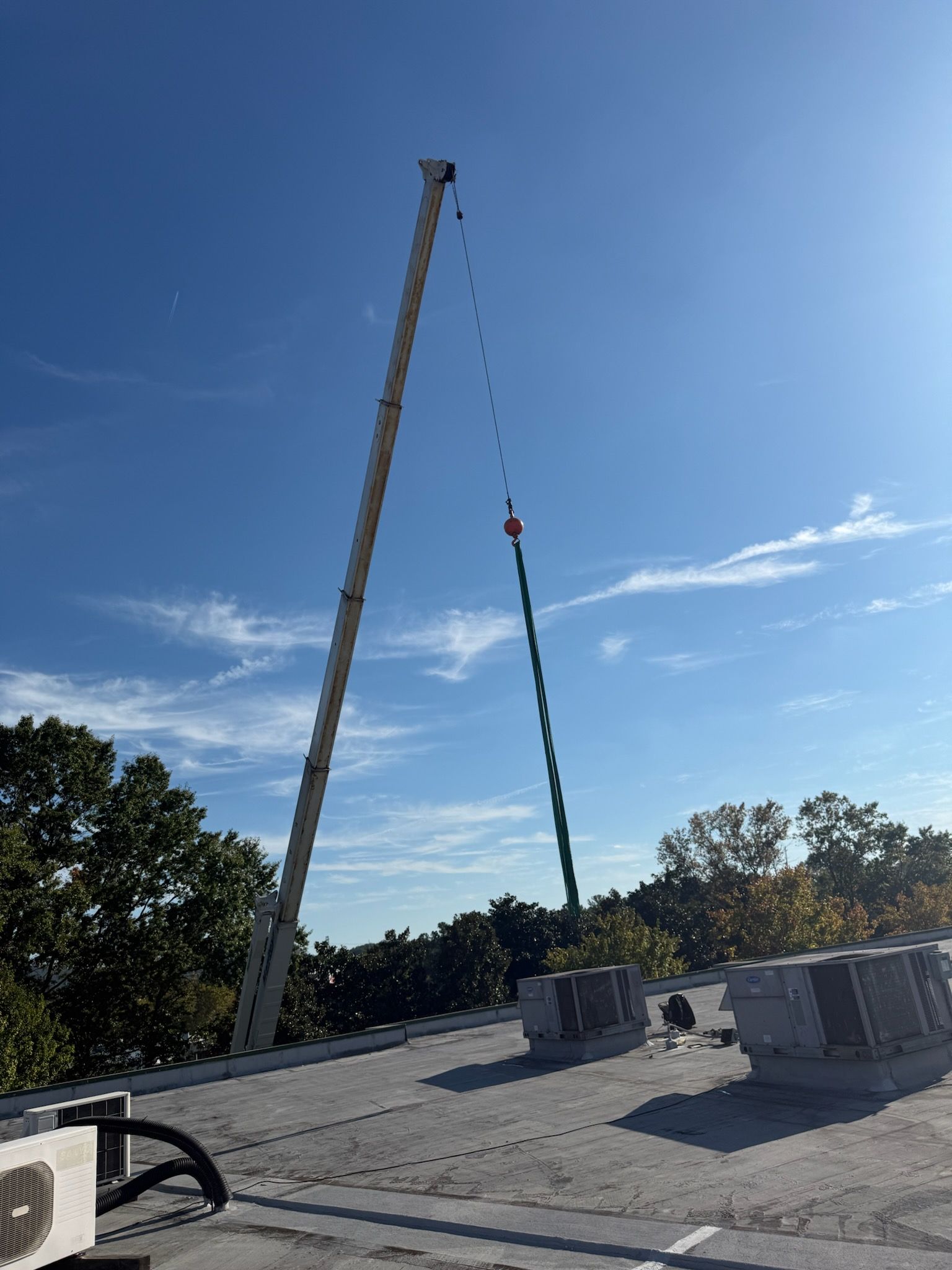 Crane lifting equipment onto a rooftop against a blue sky, trees in the background.