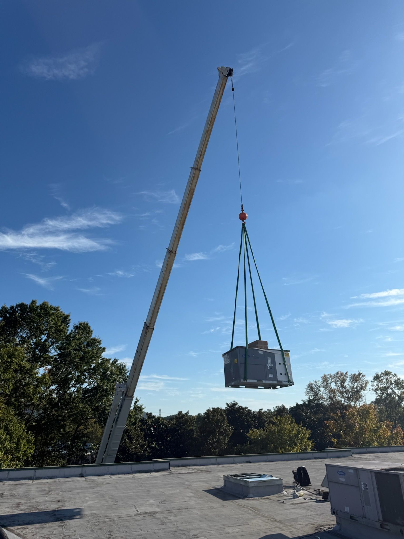 Crane lifting HVAC unit onto a rooftop. Blue sky with trees in the background.