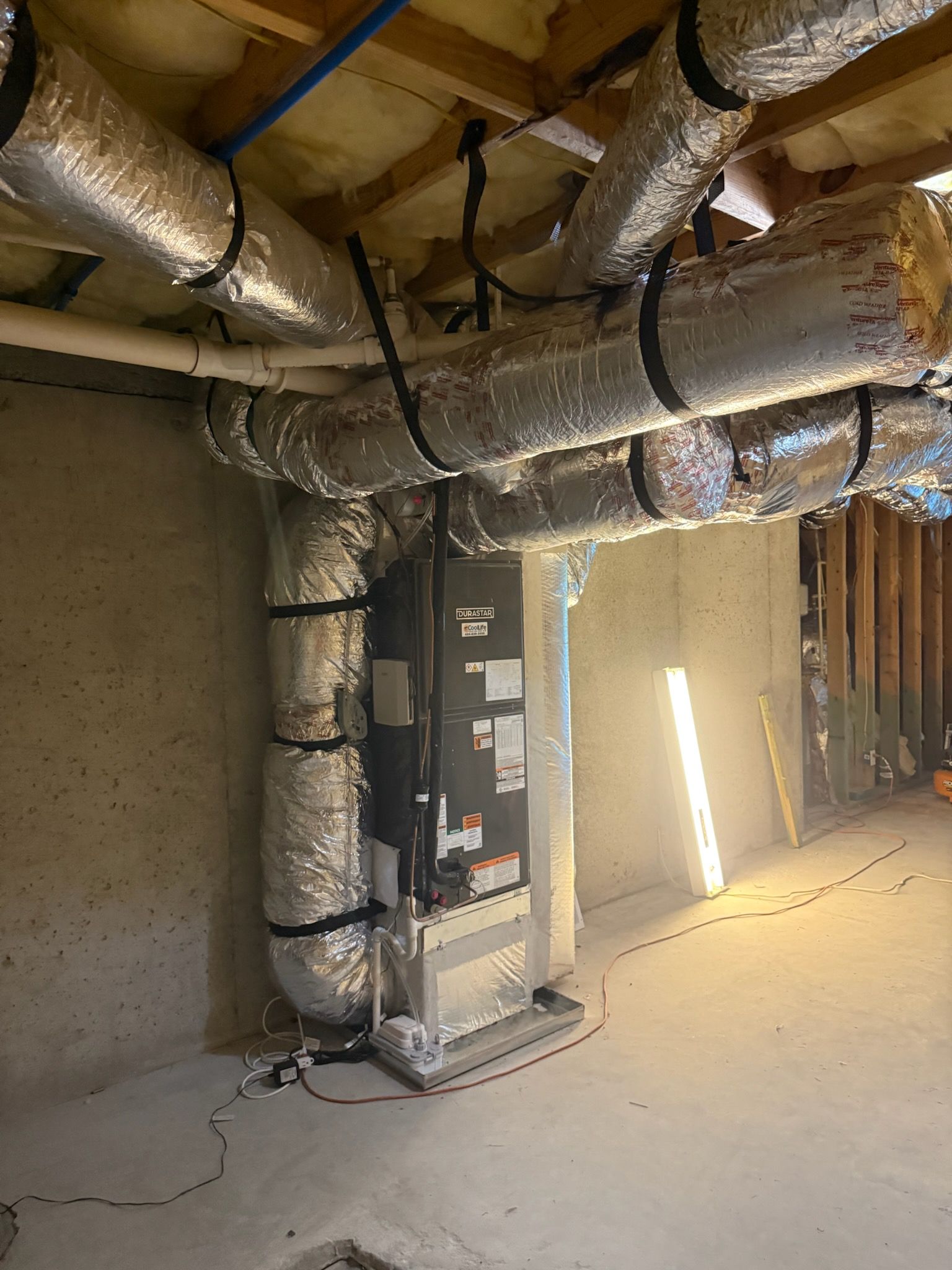 HVAC system with silver insulated ducts in a basement. The unit is tall and rectangular.
