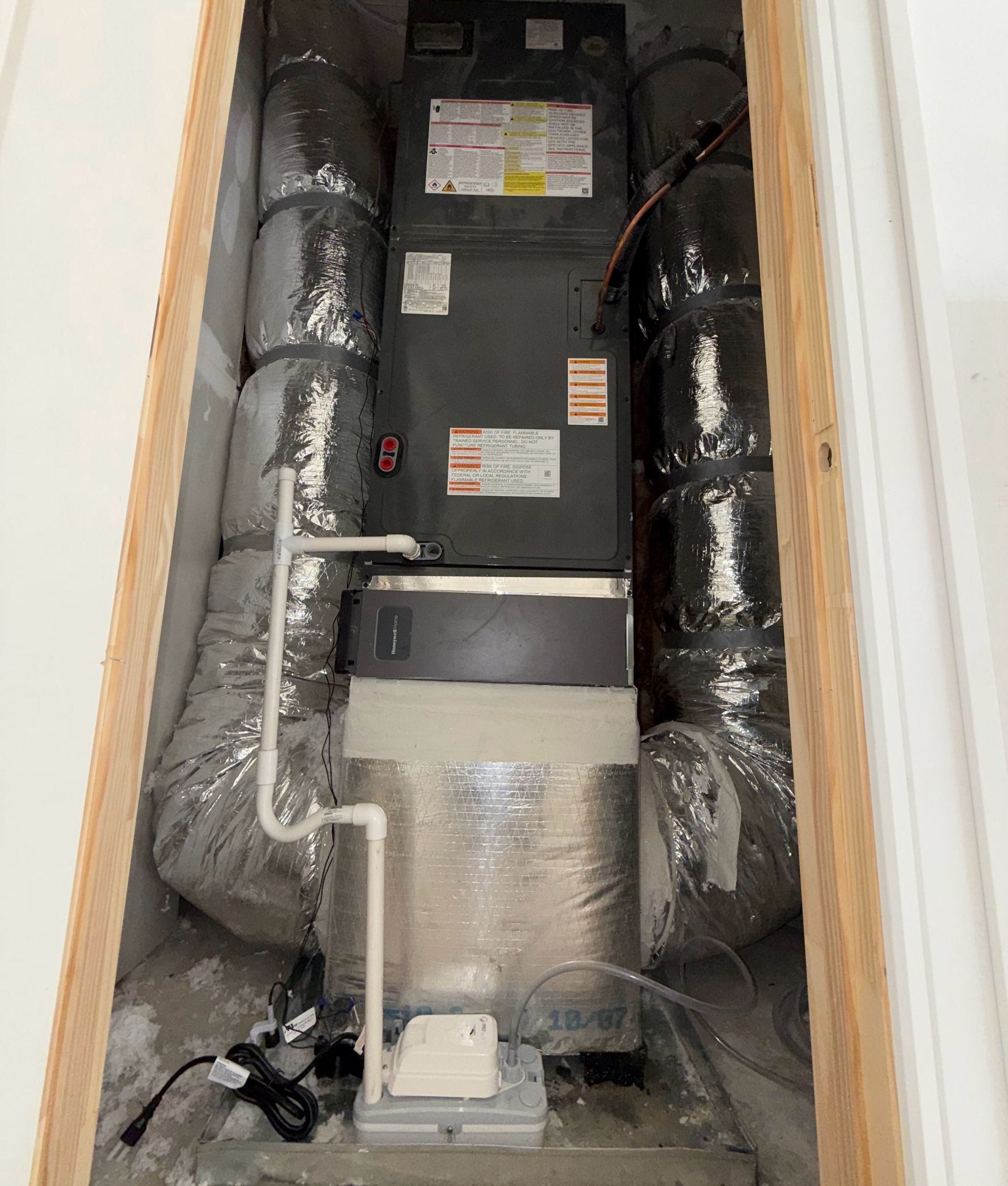 HVAC unit with ductwork in an enclosed space. Silver ducts, white pipes, and a grey unit are visible.