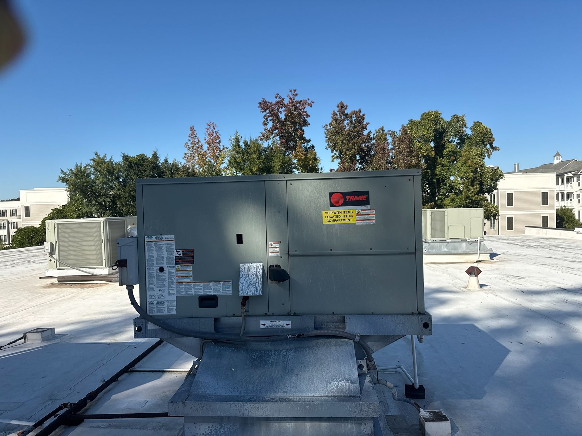 HVAC unit on a rooftop, sunny day. Gray unit, white roof, trees and buildings in background.