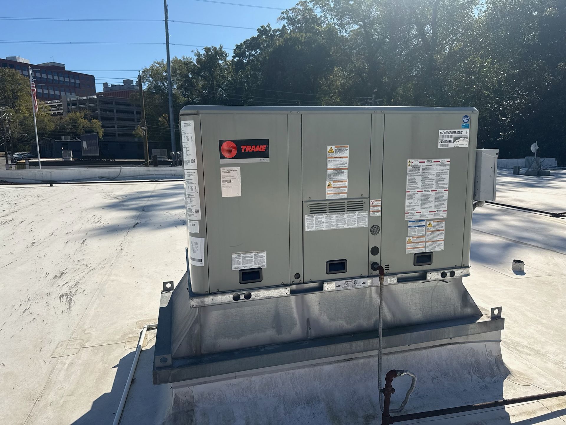 Gray Trane HVAC unit on a white roof on a sunny day. Buildings and trees are in the background.