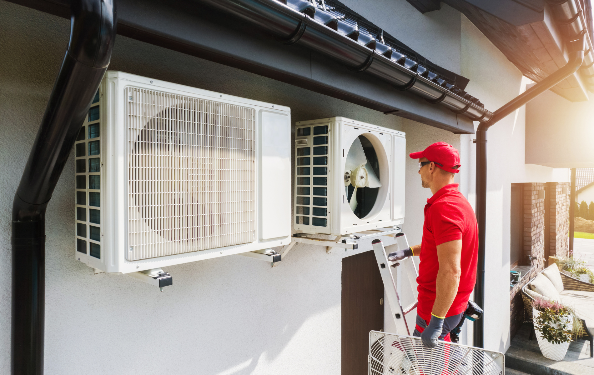 HVAC technician in red shirt installing air conditioning units on a house, sunny day.