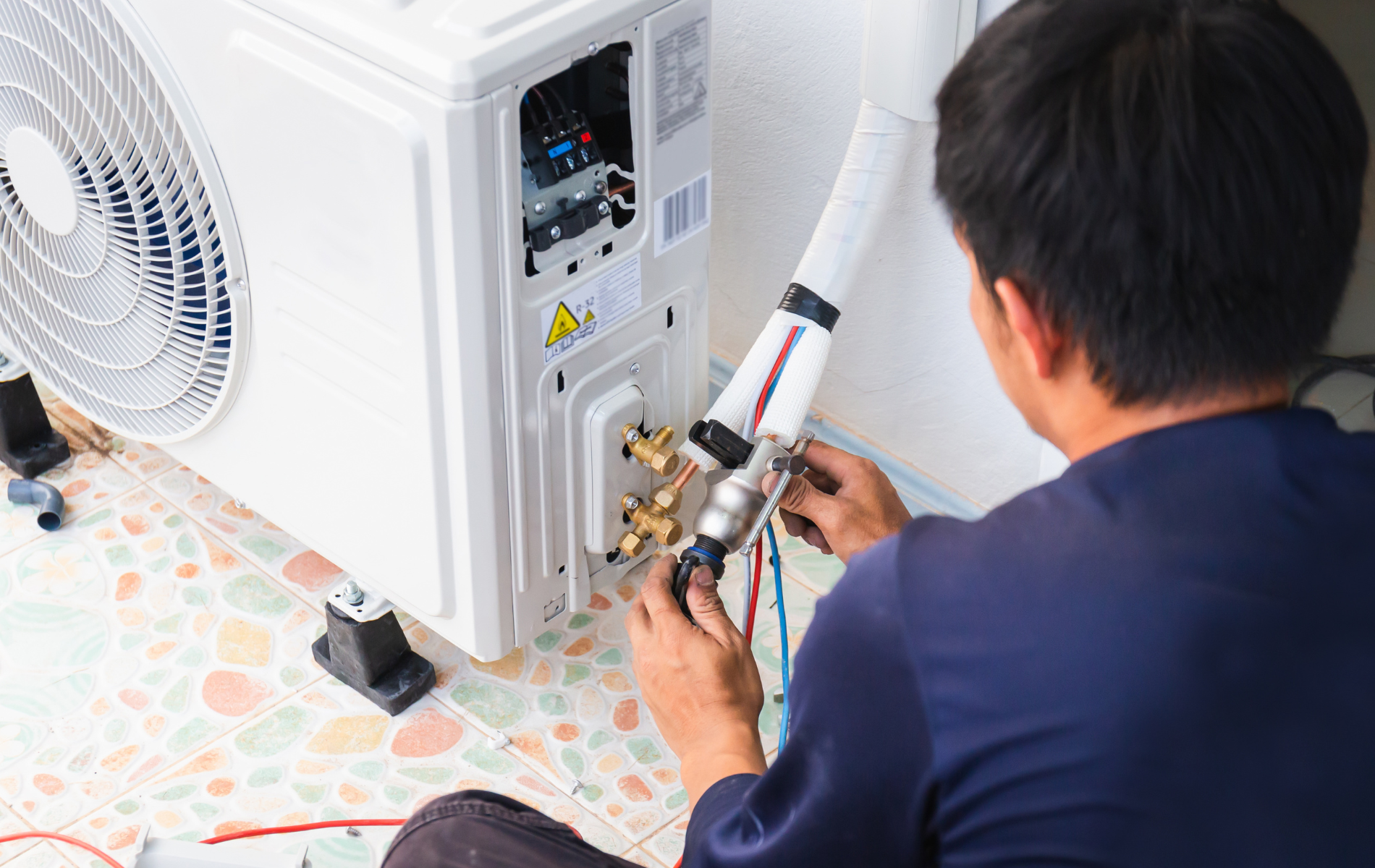 Man connecting pipes to an air conditioning unit outside on a tiled floor.