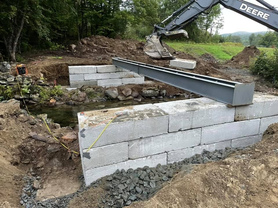 A deere excavator is lifting a beam over a concrete wall.