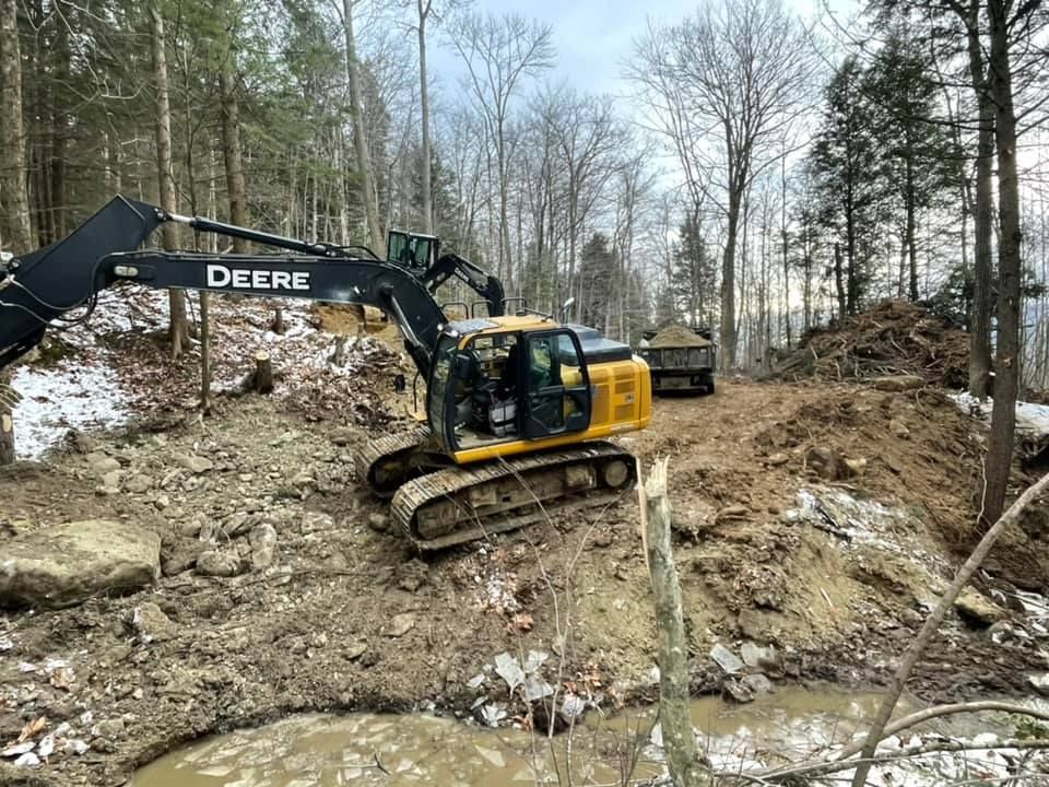 A yellow and black excavator is moving dirt in the woods.
