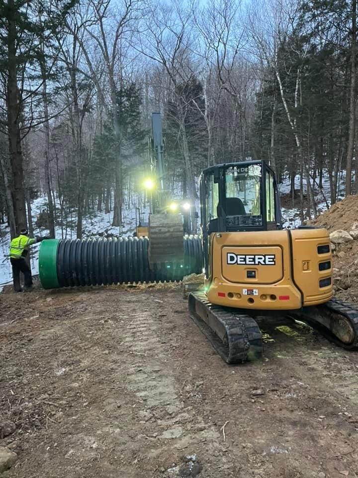 A deere excavator is working on a pipe in the woods.