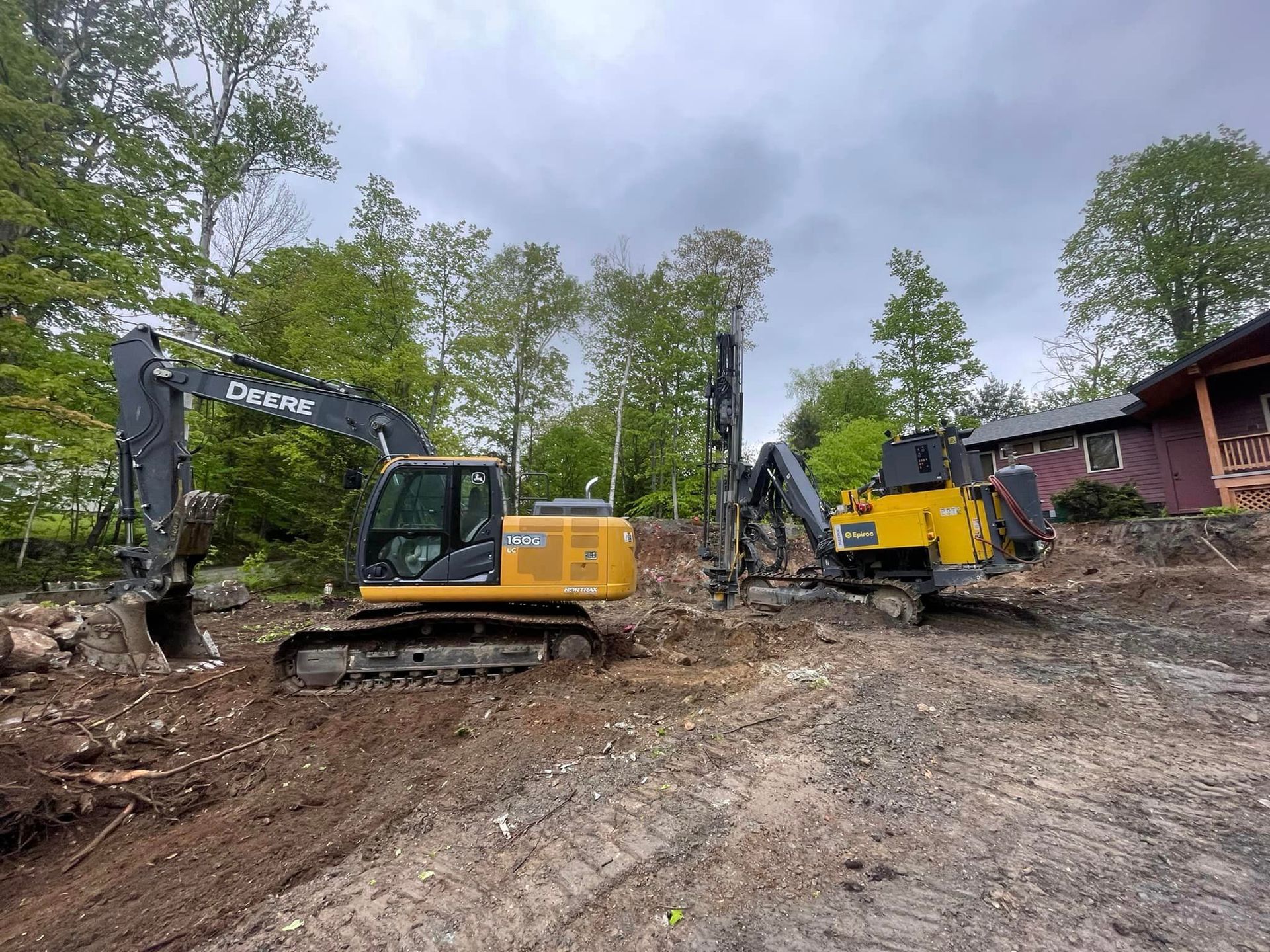 Two excavators are working on a dirt field in front of a house.