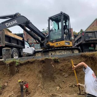 A man is digging a hole in front of a deere excavator.