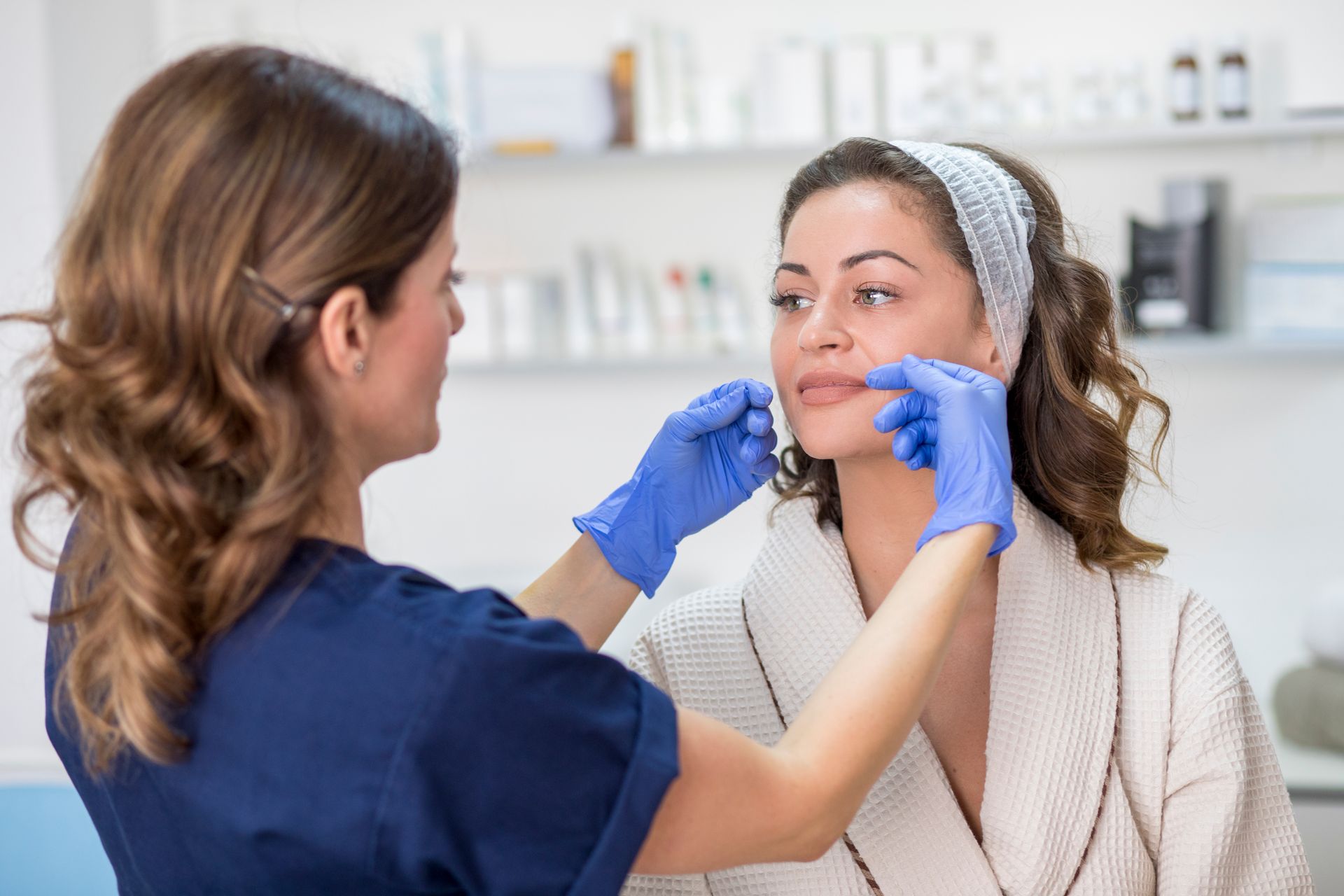 A woman in a bathrobe receives a facial assessment from a person wearing blue gloves.