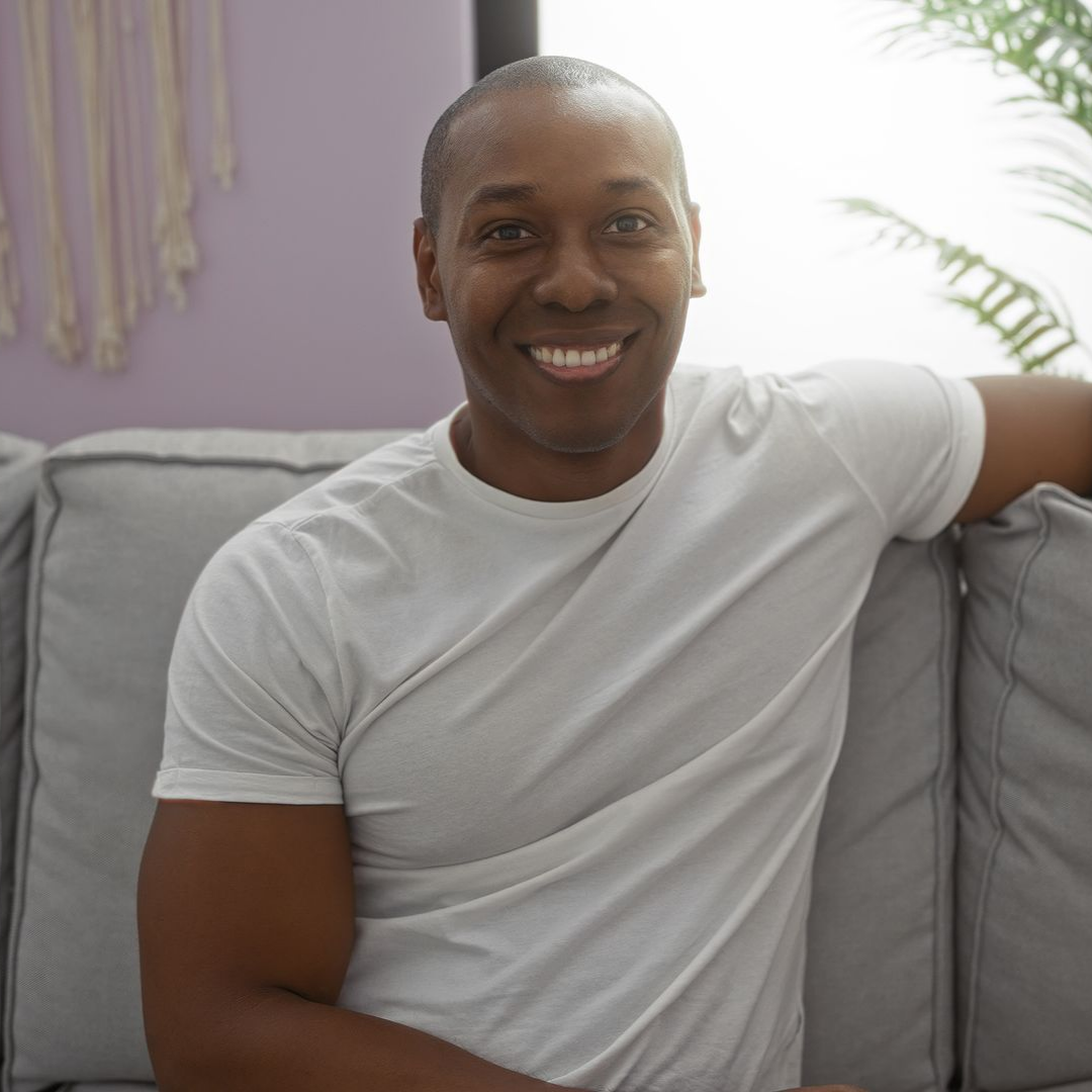 Man smiles while seated on a gray couch in a room with a light purple wall. Man smiles while seated on a gray couch in a room with a light purple wall.