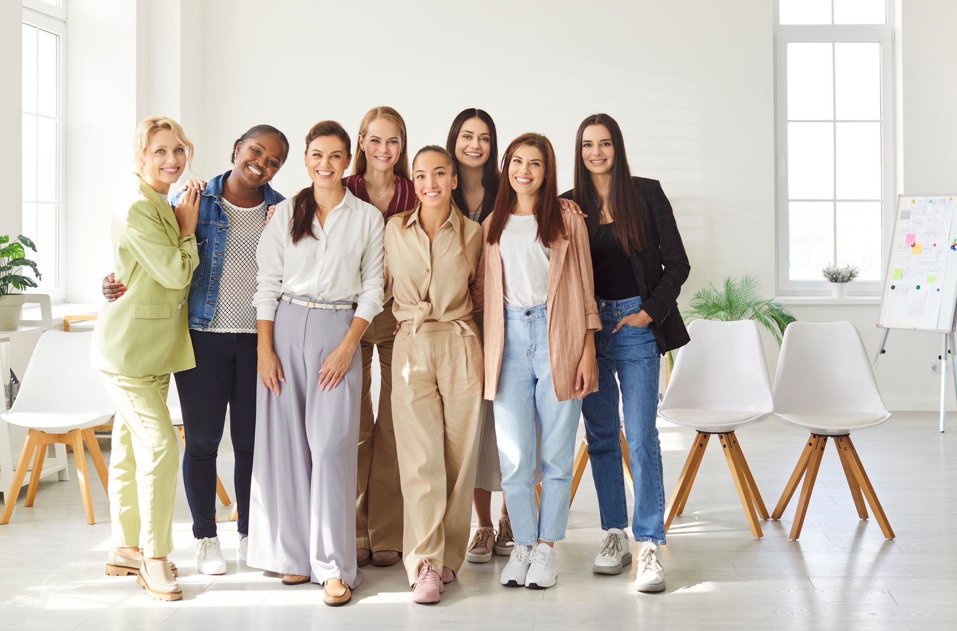 Group of women smiling together in a brightly lit room with white chairs.