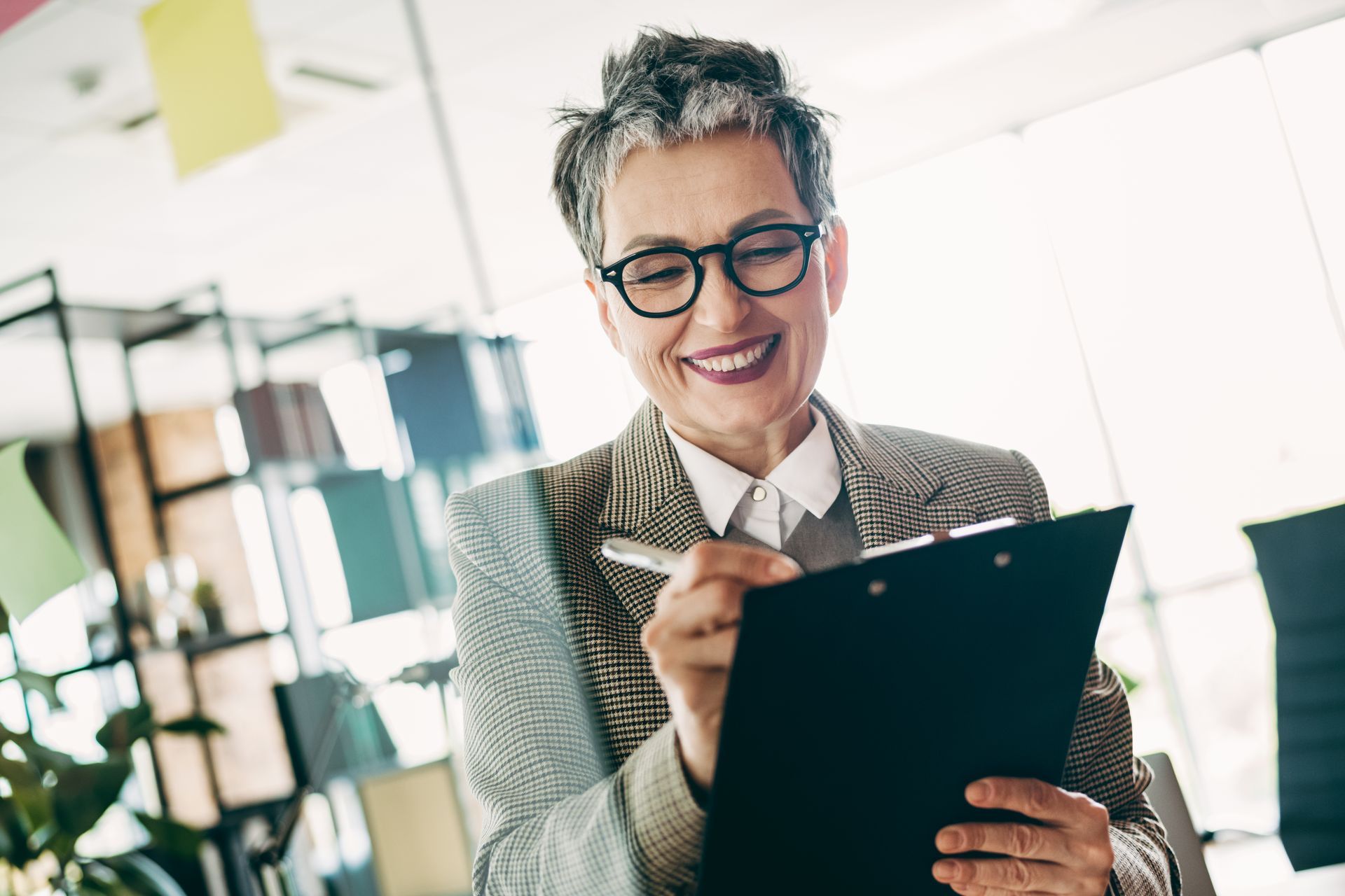 Woman with glasses smiles while writing on a clipboard in an office setting. Woman with glasses smiles while writing on a clipboard in an office setting.