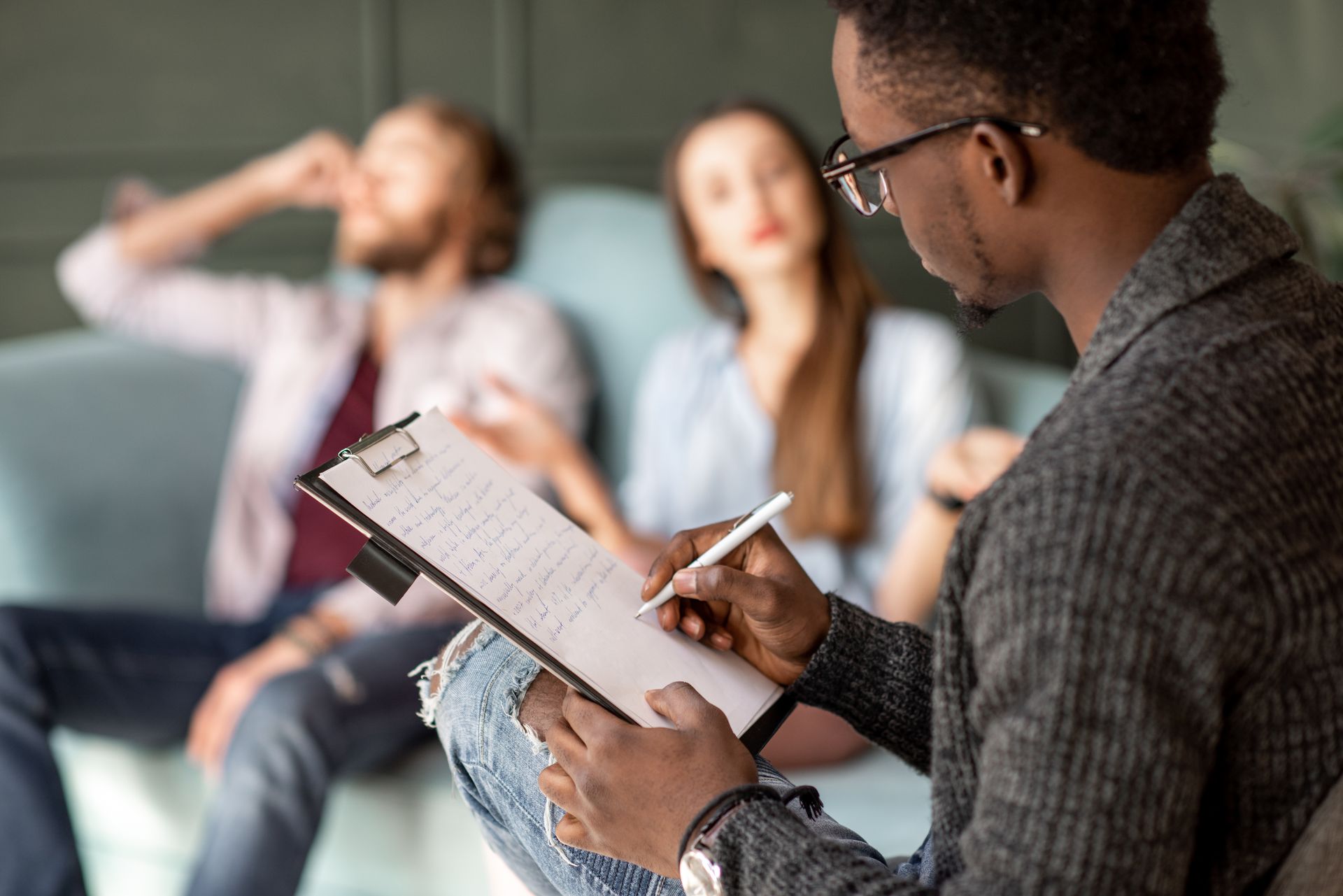 Therapist taking notes while counseling a couple. They sit on a couch in an office setting.