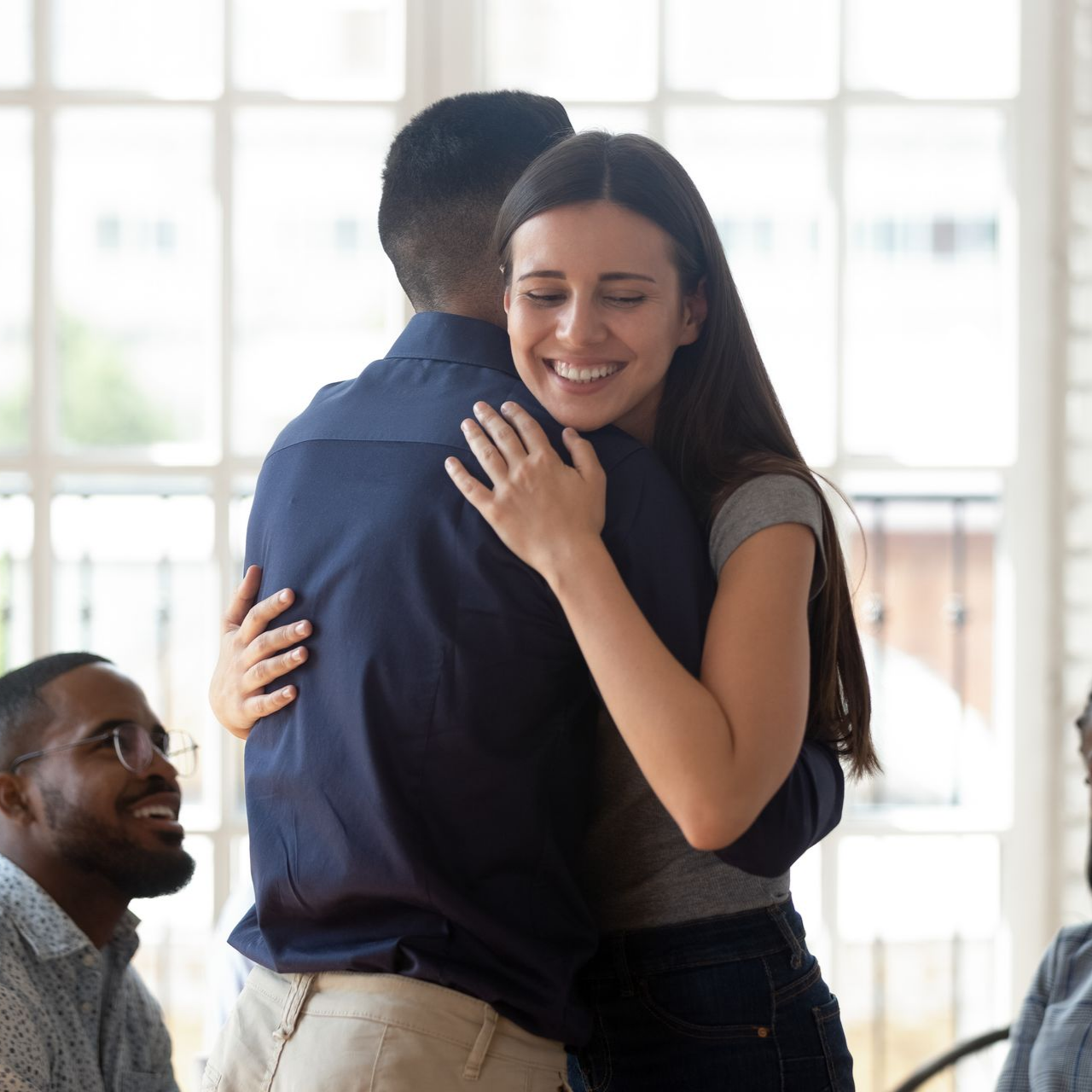 Woman hugs a person in a blue shirt indoors; others watch smiling.