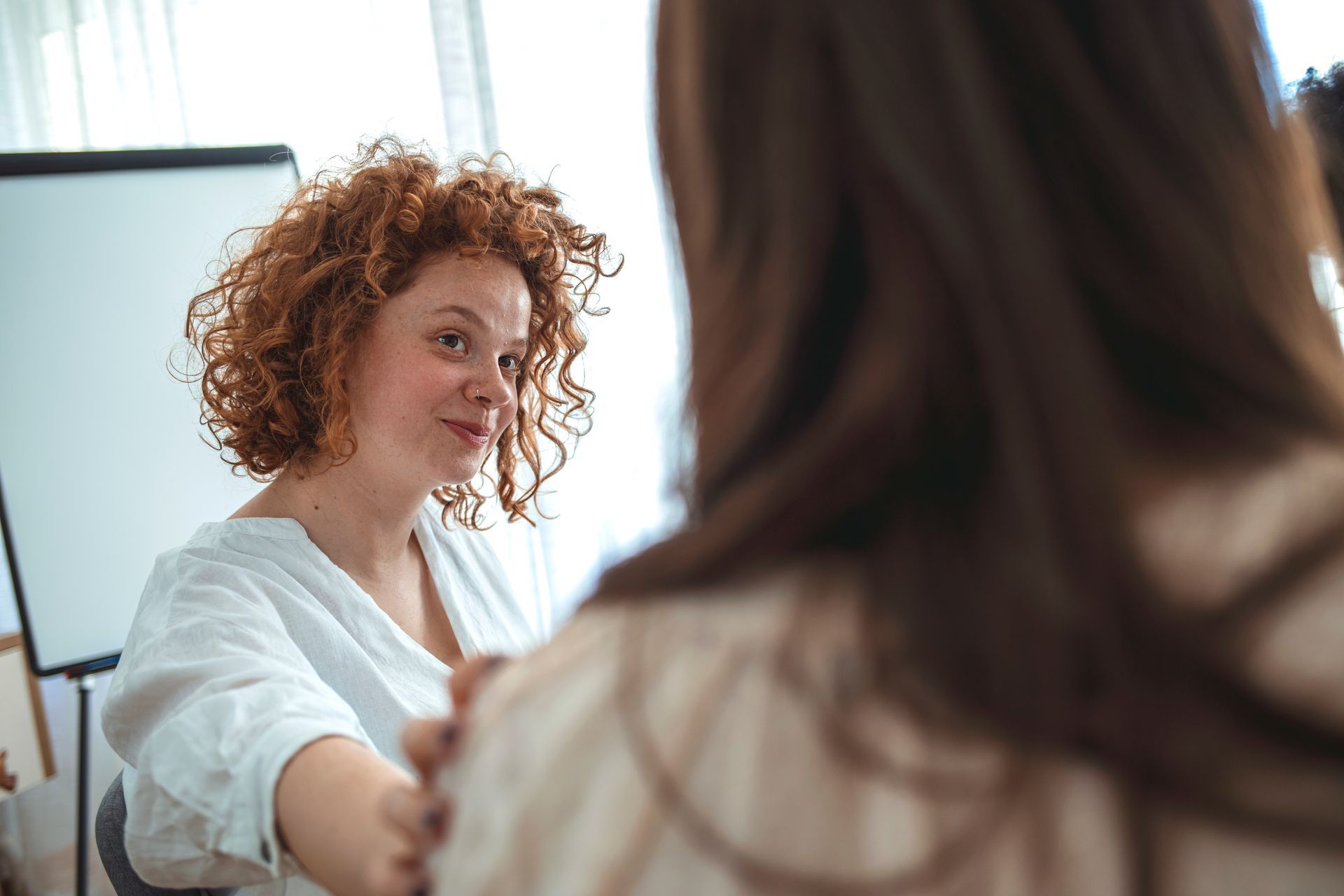 Woman with curly red hair reaches out to another woman. White backdrop and whiteboard visible.