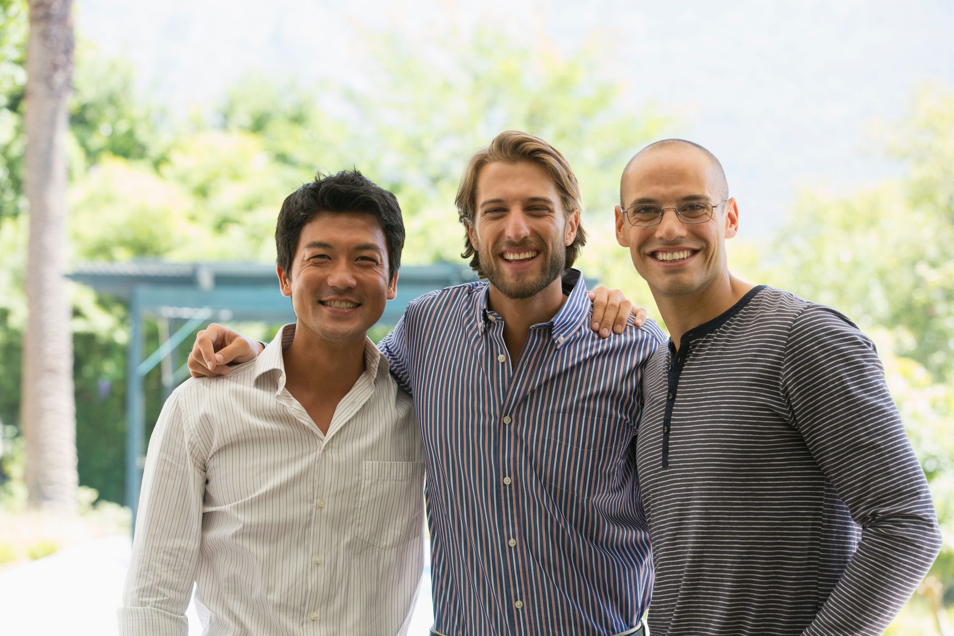 Three men smiling, arms around each other. Outdoor setting with green foliage in background.