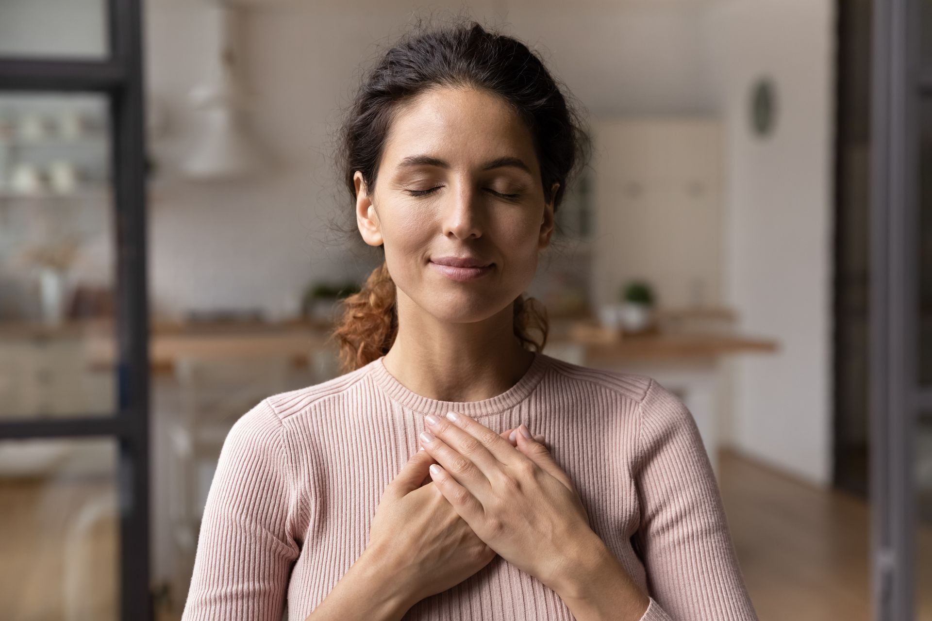 Woman with eyes closed, hands on chest, appears to meditate in a bright interior.