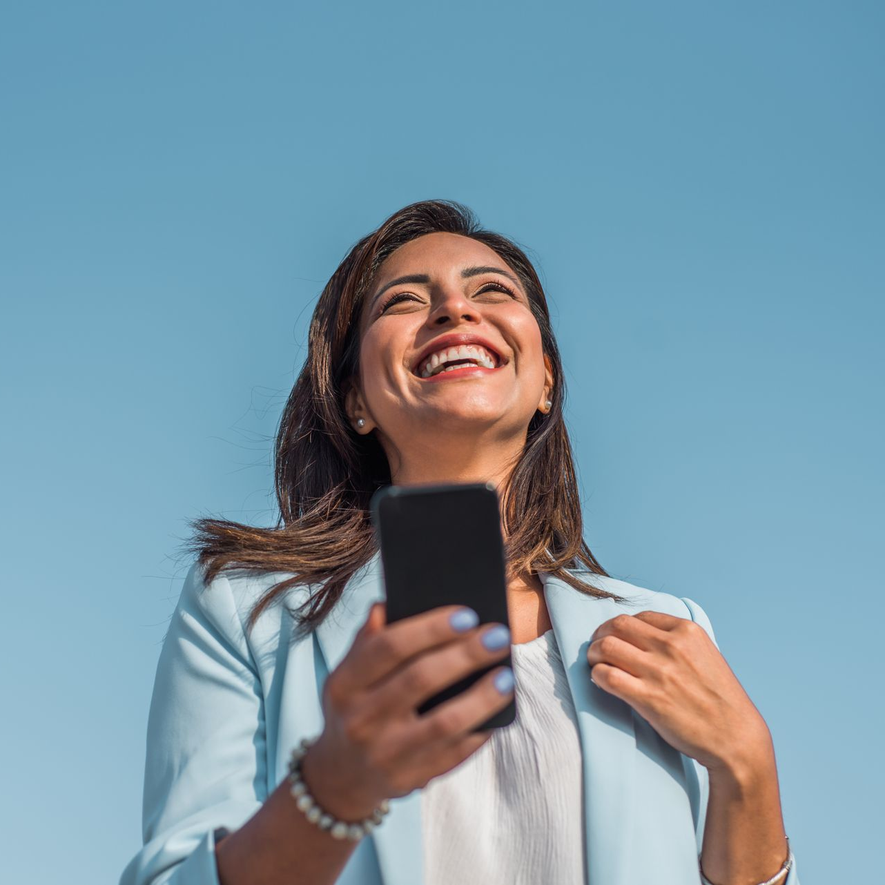 Woman in light blue blazer smiles while looking up at a blue sky, holding a black smartphone. Woman in light blue blazer smiles while looking up at a blue sky, holding a black smartphone.