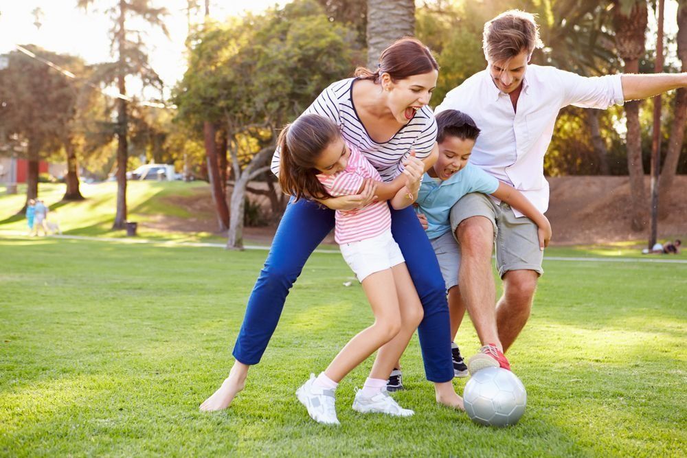 Family playing soccer together on a sunny park lawn, smiling and kicking a ball