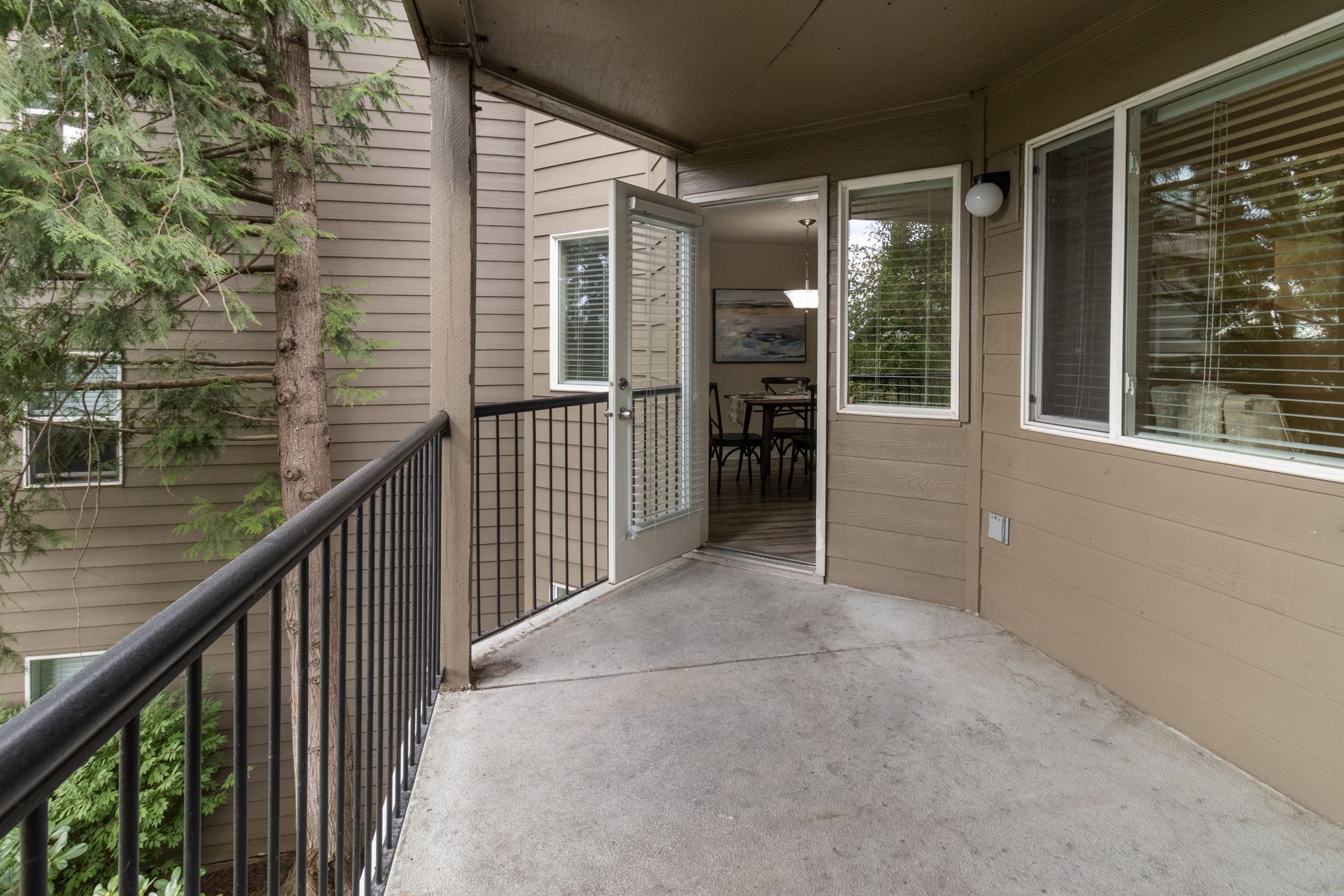 Balcony with open doors to a dining area. Black railing, concrete floor, and trees in background.