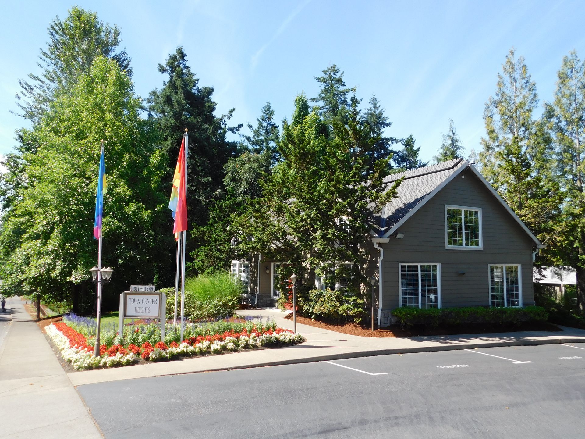 Small gray building with flags and flowerbed, trees in background, sunny day.