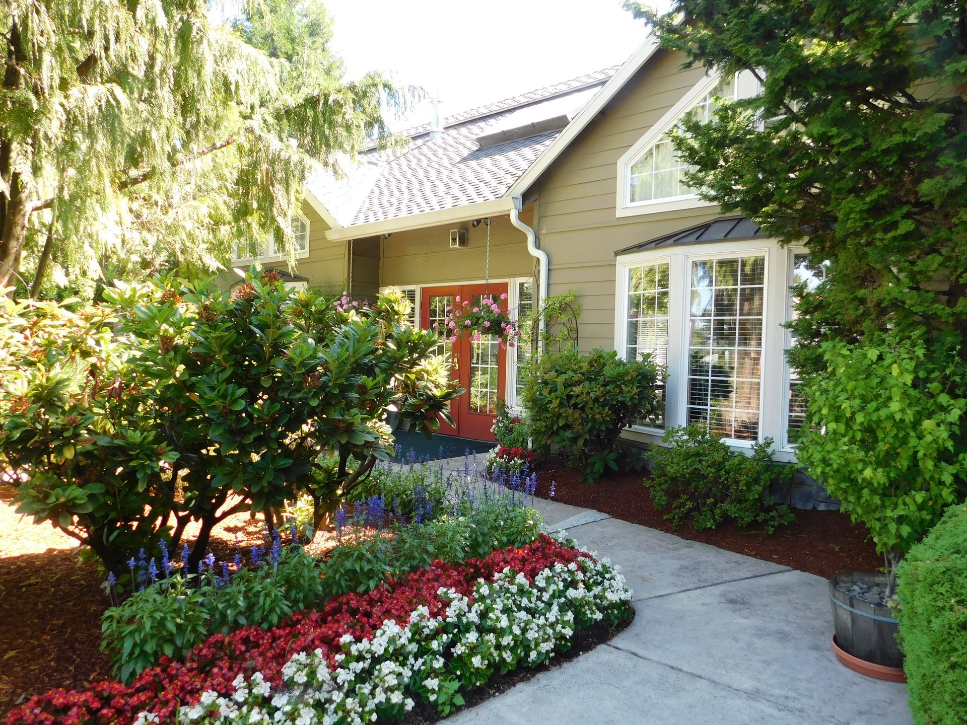 House with red door and white-trimmed windows. Flowerbeds line a walkway, framed by greenery.