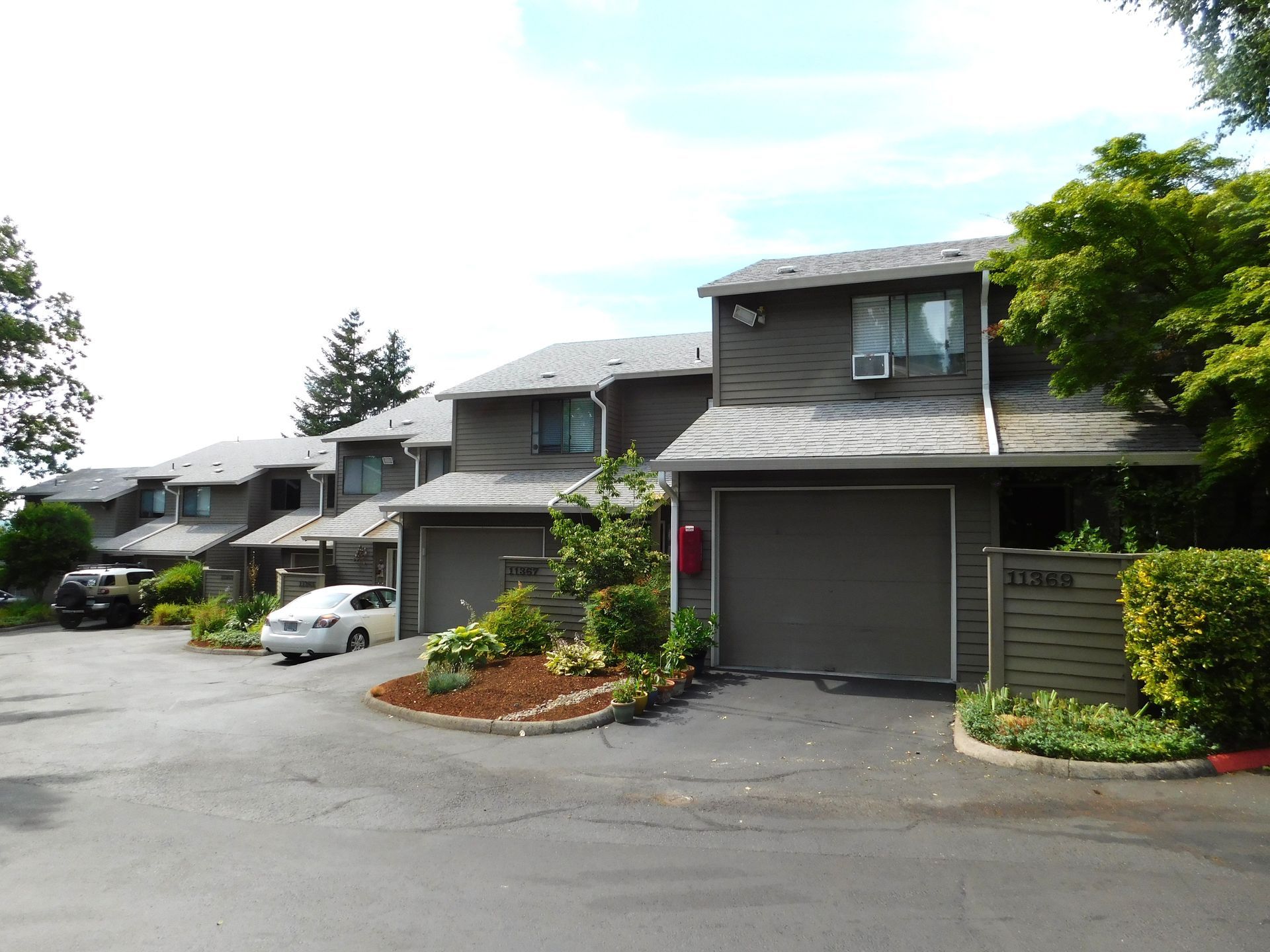 Townhomes with gray siding, driveways, and garages. Green trees and shrubs surround the buildings.
