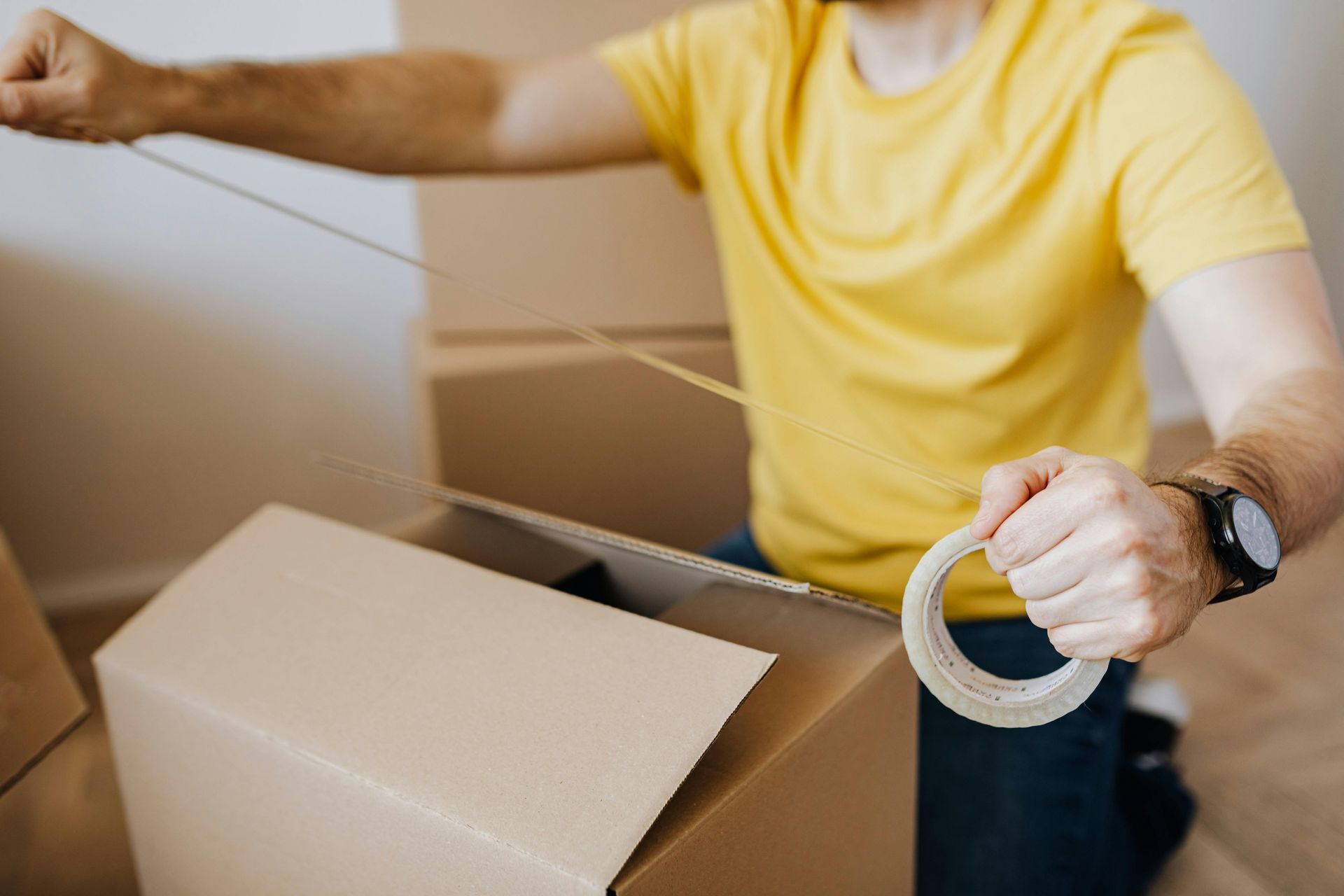 Person in a yellow shirt sealing a cardboard box with packing tape, preparing for moving or packing items