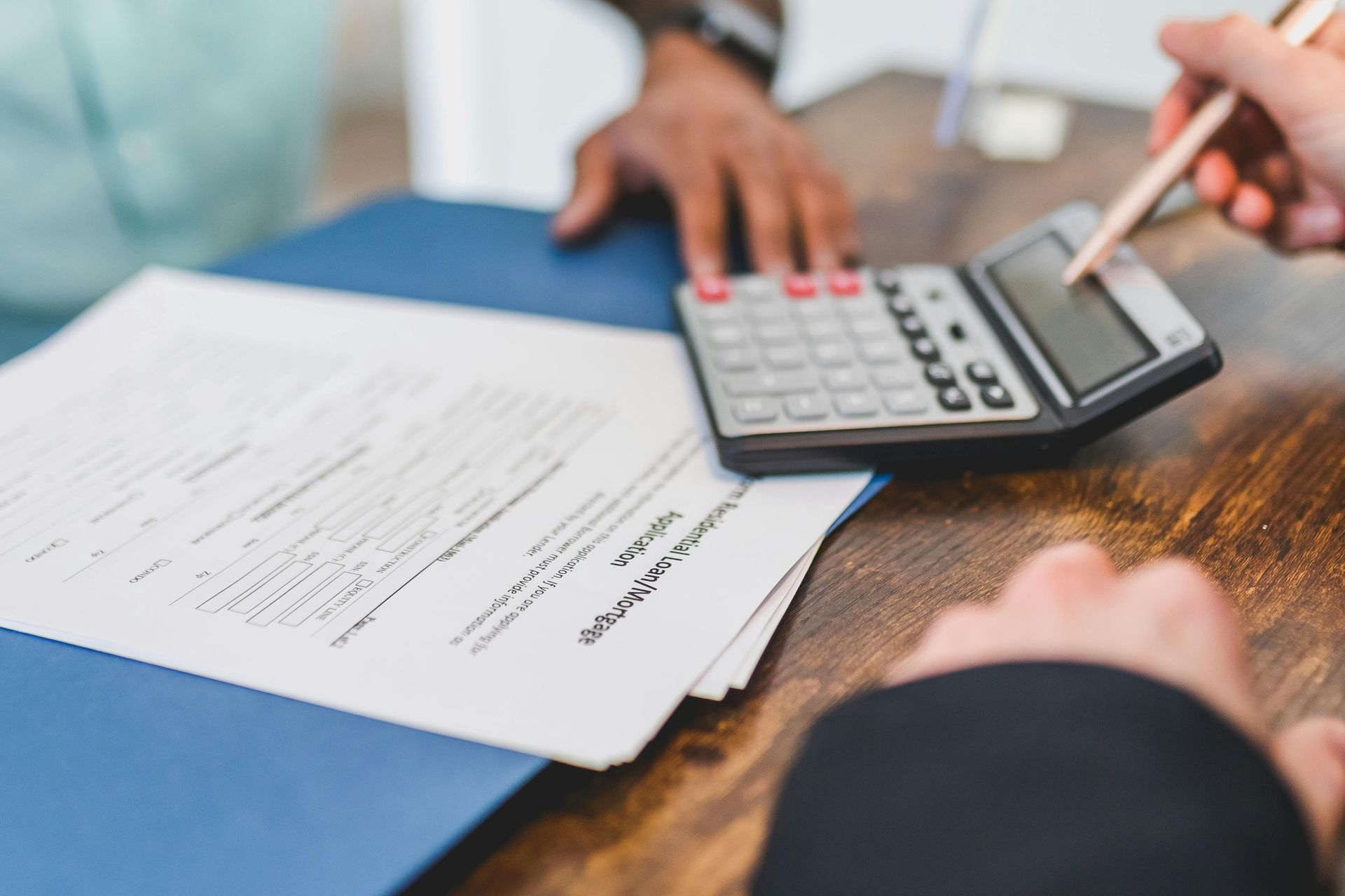 Close-up of two people working on financial documents and using a calculator. One person is pointing at the calculator with a pen, while a loan application form is visible on the table