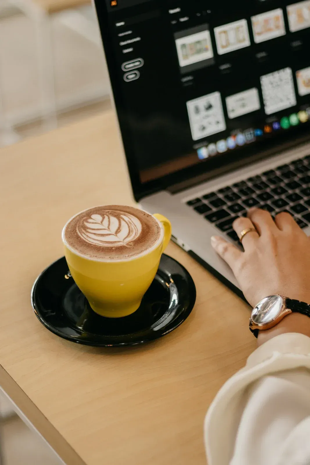 Laptop on table next to yellow coffee cup and black saucer, hand typing on keyboard.