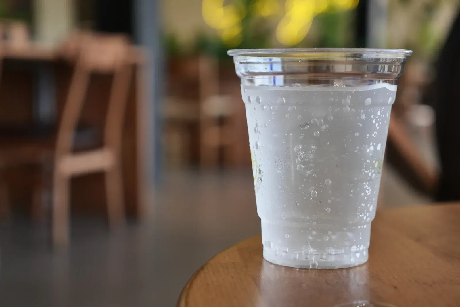 Clear plastic cup with condensation, filled with water, on a wooden table. Blurred café background.