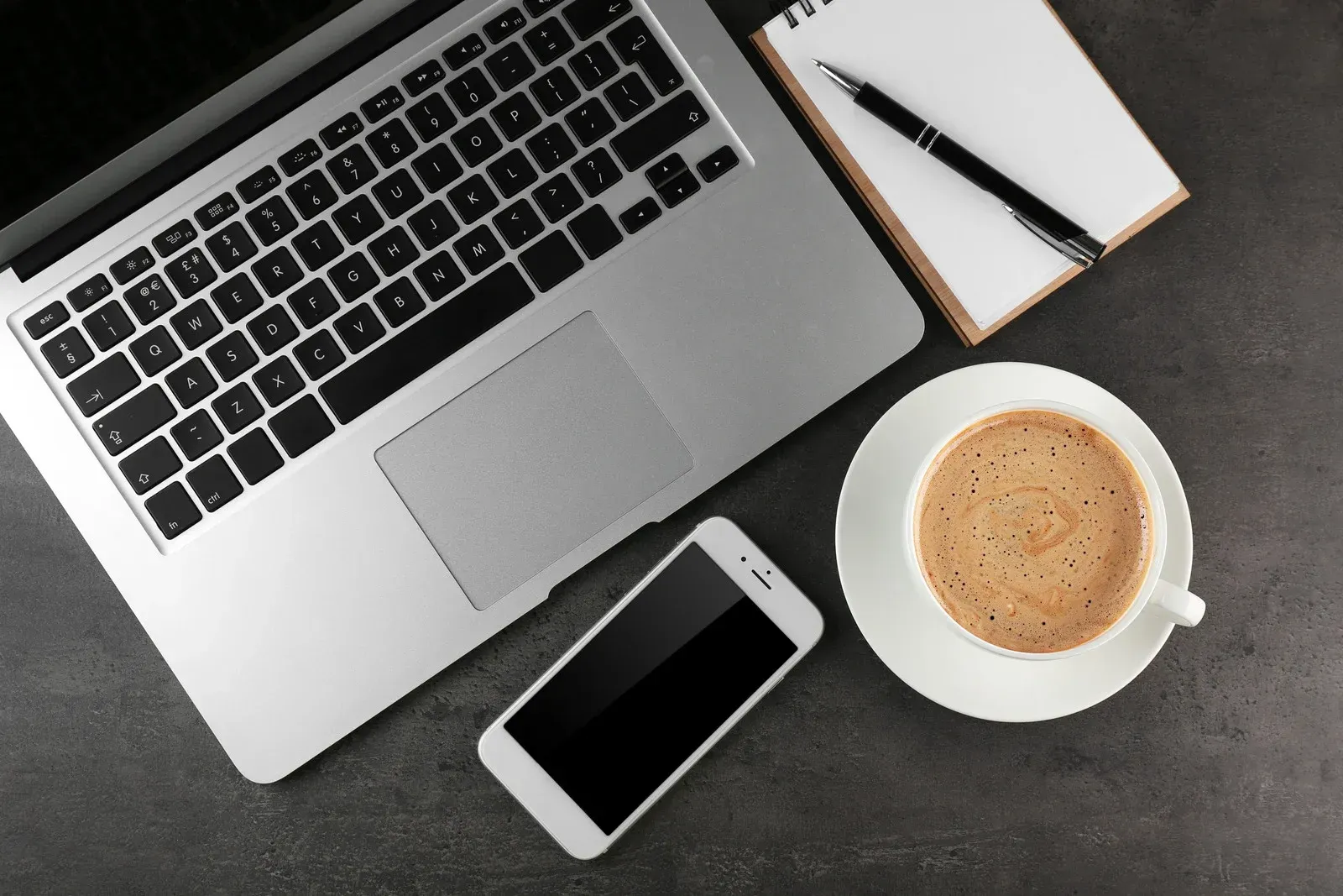 Laptop, phone, notepad with pen, and coffee cup on a gray surface.