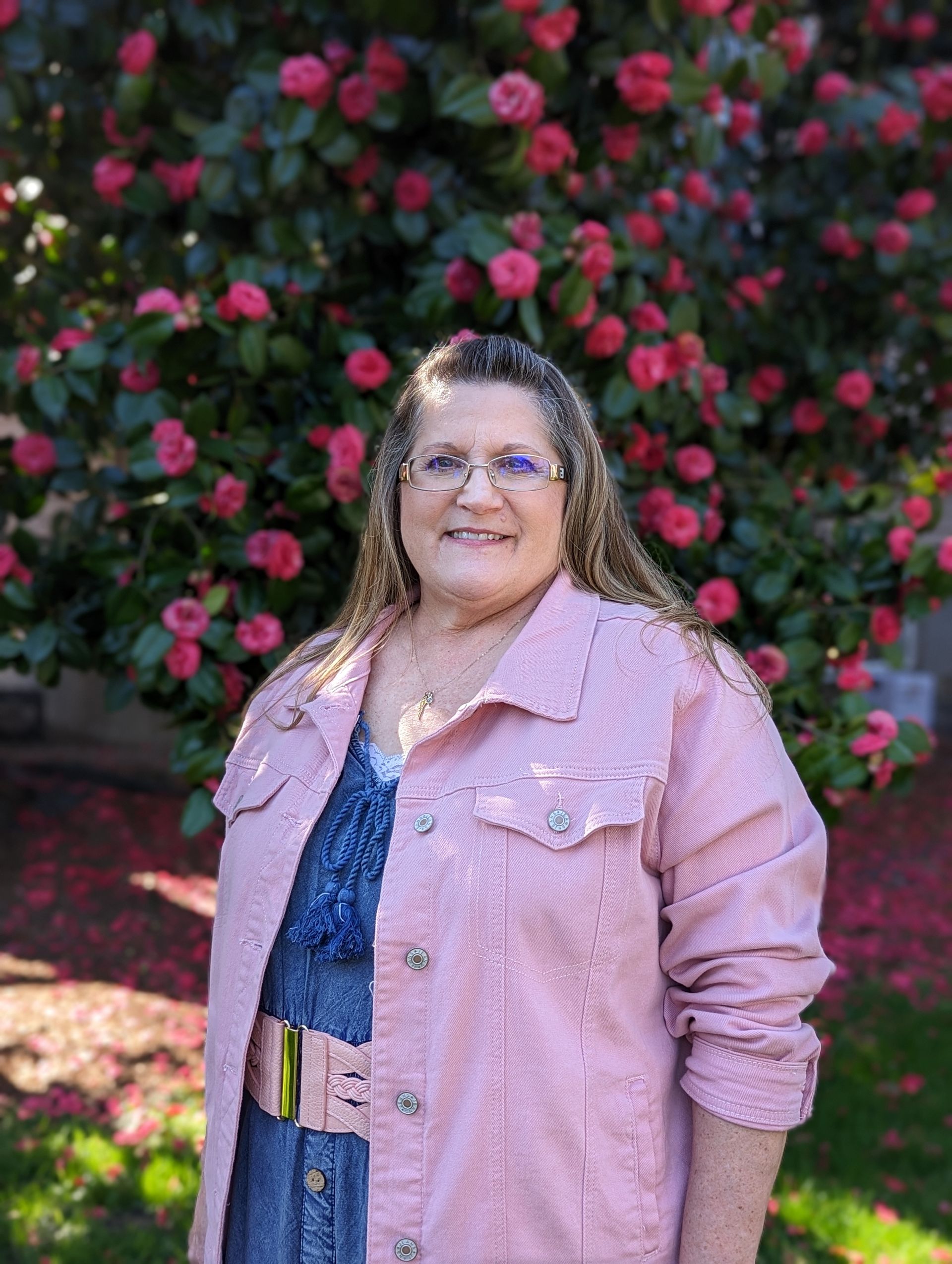 A woman in a pink jacket and glasses is standing in front of a tree with pink flowers.