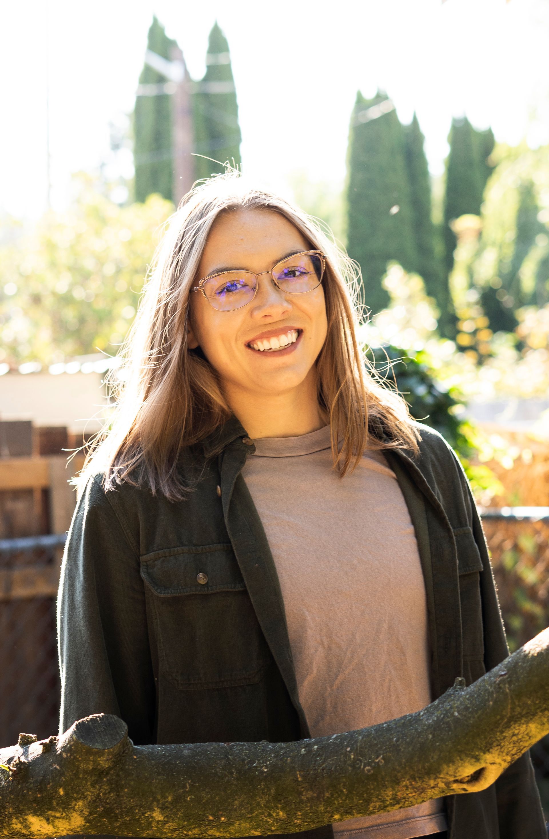 A woman wearing glasses is standing next to a tree branch and smiling.