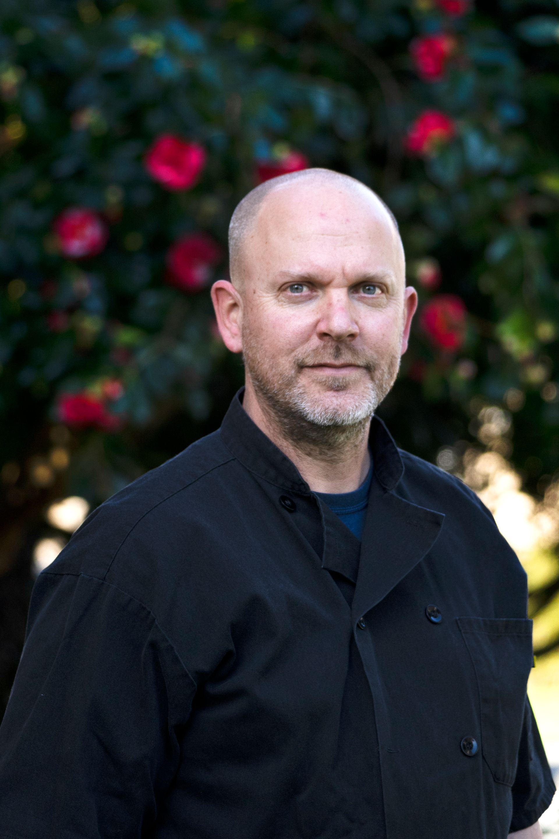 A bald man with a beard is standing in front of a tree with red flowers.