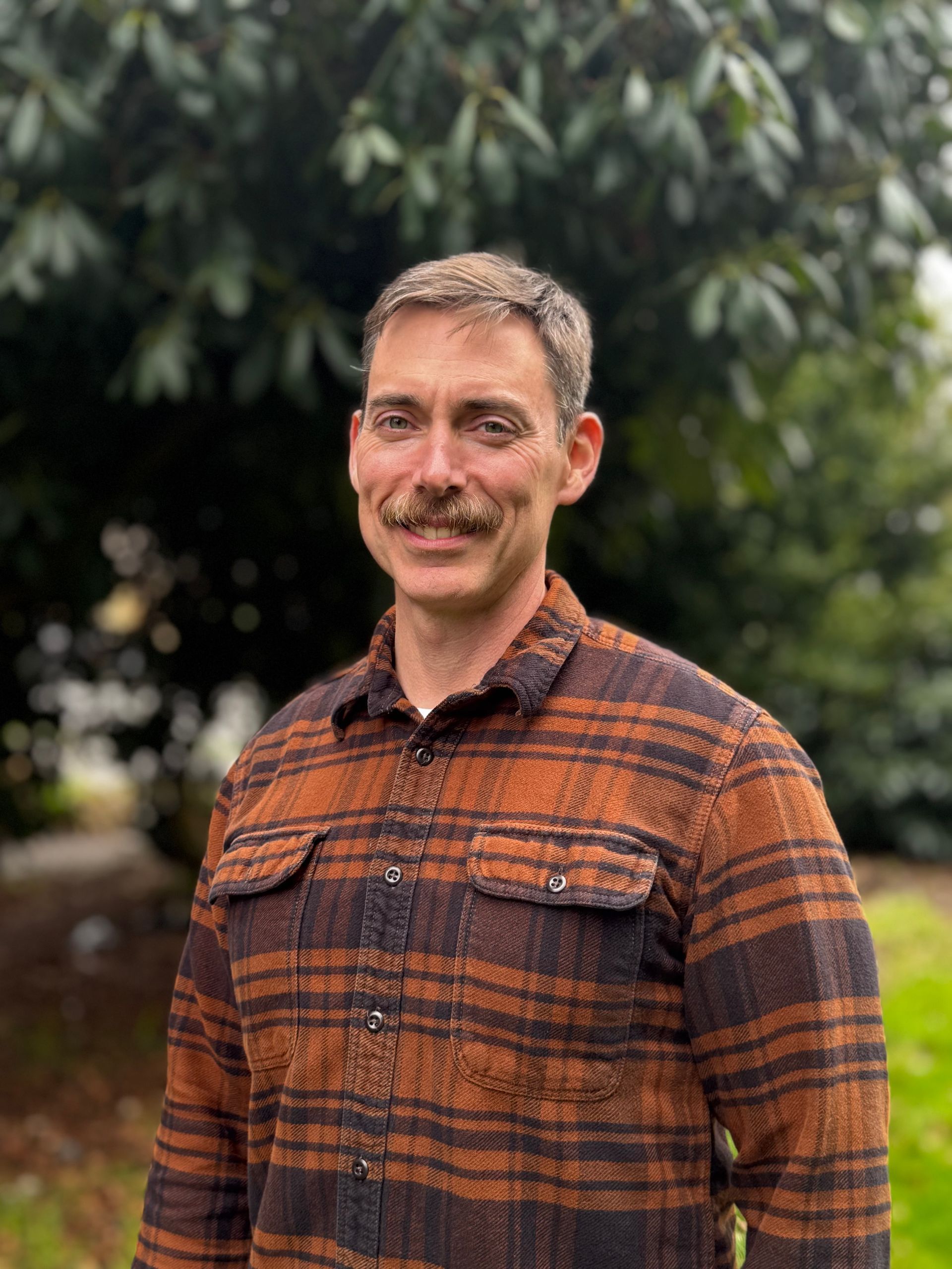 A man in an orange/brown shirt is standing in front of a tree.