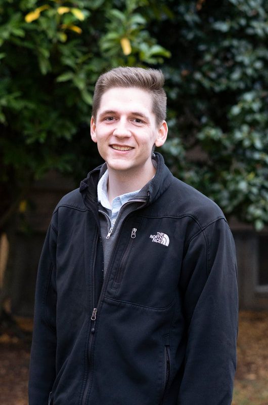 A young man wearing a black jacket is standing in front of a tree.