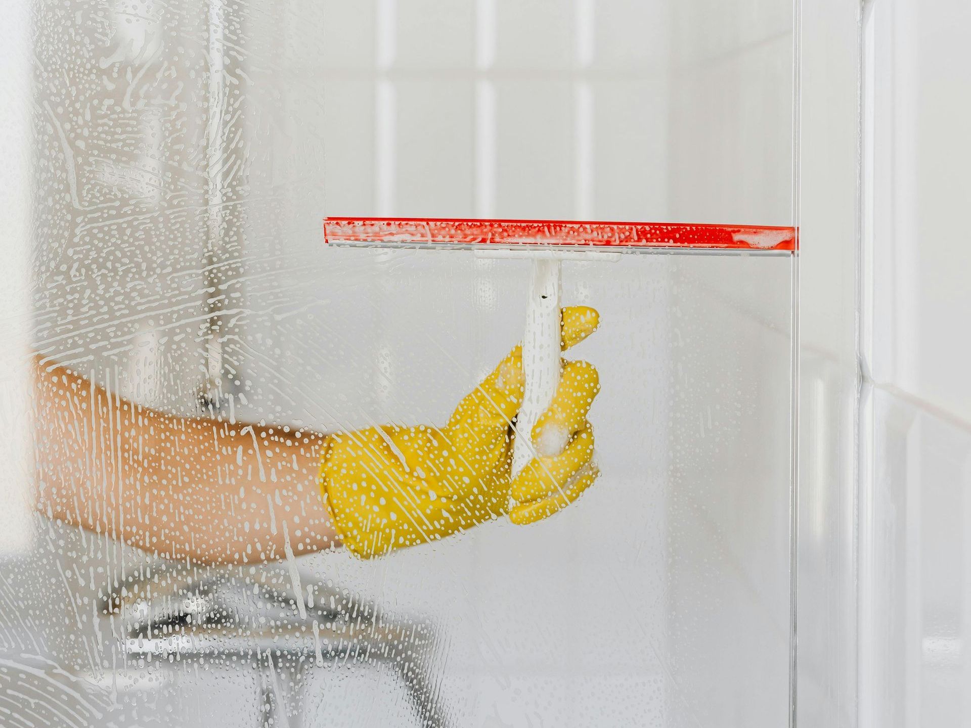 A person wearing yellow gloves is cleaning a shower door with a squeegee.