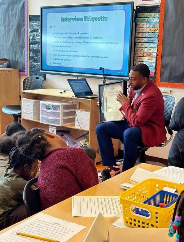 A man in a red jacket is speaking into a microphone in front of a screen that says interview etiquette