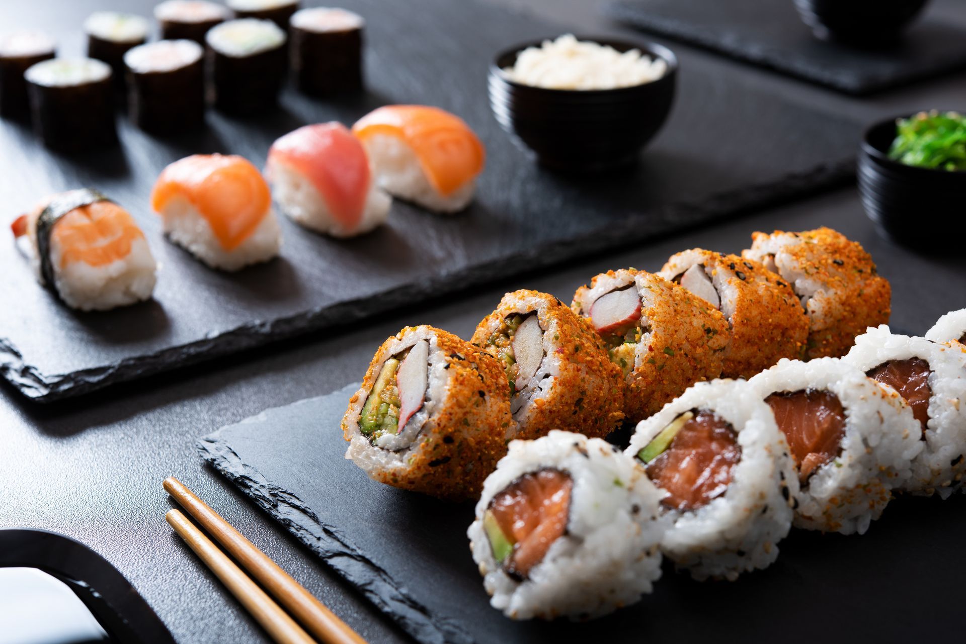 Disposizione di sushi su vassoi di ardesia nera; include nigiri, maki roll e contorni.