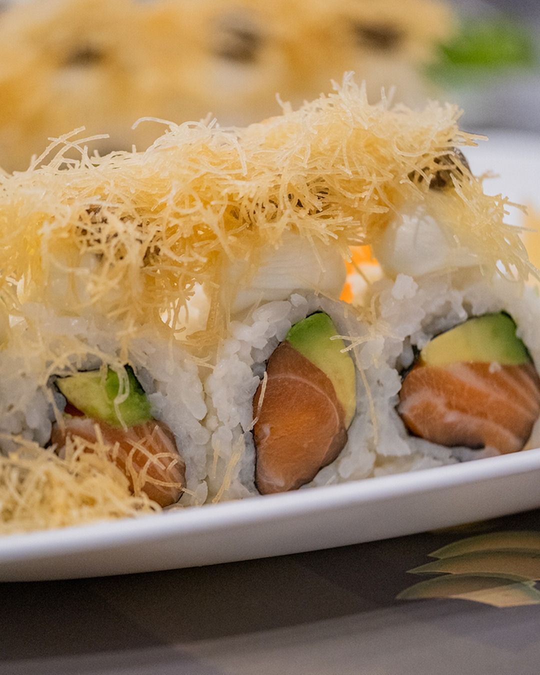 Involtini di sushi con salmone, avocado e guarnizione croccante su un piatto bianco.